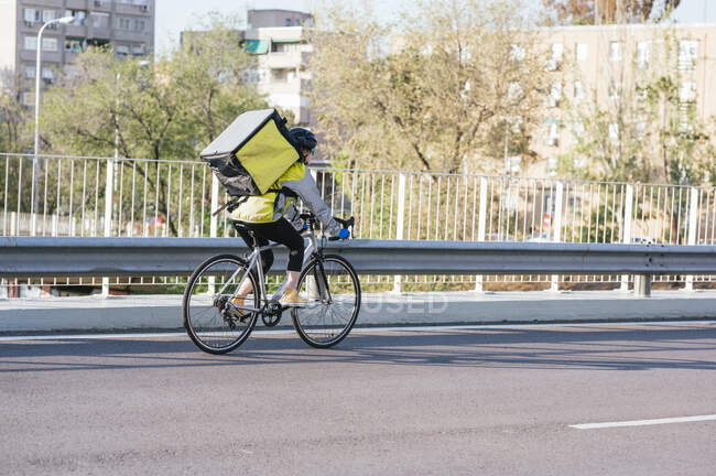 Back view of female courier with thermal bag riding bike on bridge while delivering food in city — Stock Photo