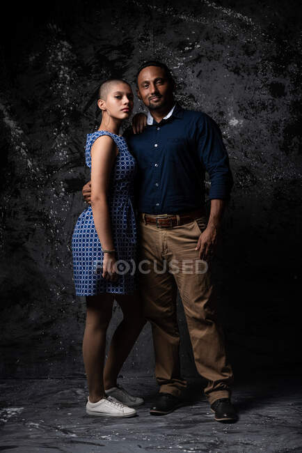 Tender ethnic man embracing woman on dark background in studio looking at camera — Stock Photo