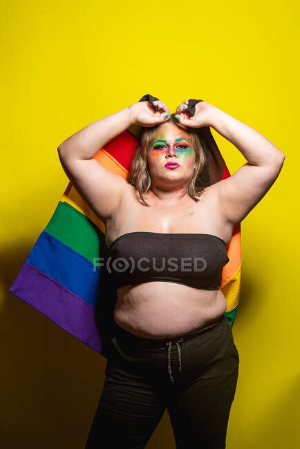 Избыточный вес женской модели с творческим макияжем, показывающий флаг ЛГБТ и смотрящий на камеру на желтом фоне — стоковое фото