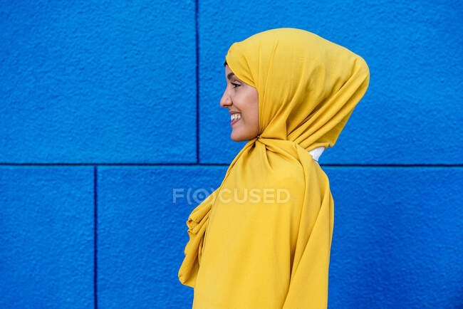 Vista laterale della donna musulmana alla moda in hijab giallo in piedi guardando lontano in strada su sfondo blu — Foto stock