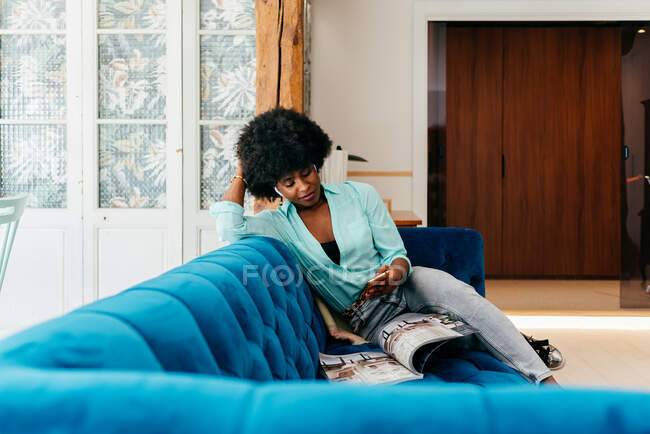 Linda jovem afro-americana em roupas casuais navegando smartphone e revista de leitura enquanto descansa no sofá azul aconchegante em casa — Fotografia de Stock