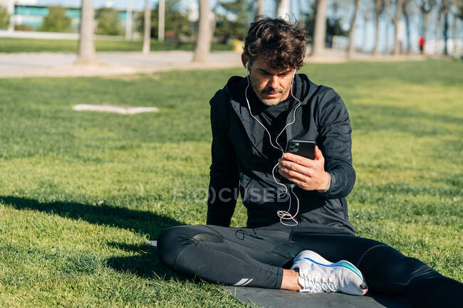 Bearded male athlete in sportswear and earphones text messaging on cellphone while sitting on meadow in town — Stock Photo