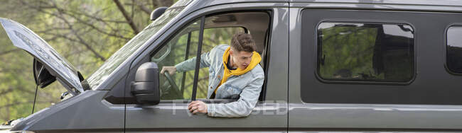Amplio ángulo de chico joven en ropa casual asomándose por la ventana de la furgoneta y mirando hacia otro lado mientras intenta iniciar el vehículo en reparación durante el goteo en la naturaleza - foto de stock
