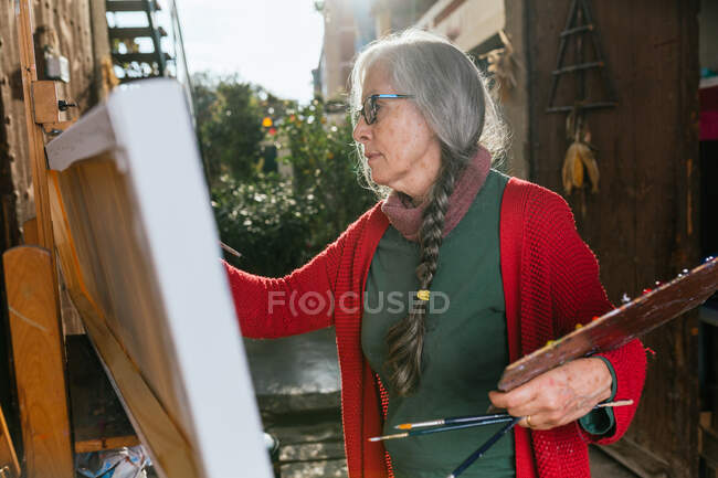 Vista lateral de artistas femeninas envejecidas en pintura de máscara protectora sobre lienzo en el patio trasero en el día soleado en verano - foto de stock