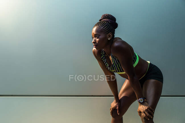 Бічний вид красивої афро-американської жінки в спорті, яка спирається на коліна і відпочиває, стоячи біля сучасної будівлі стіни на вулицях міста. — стокове фото
