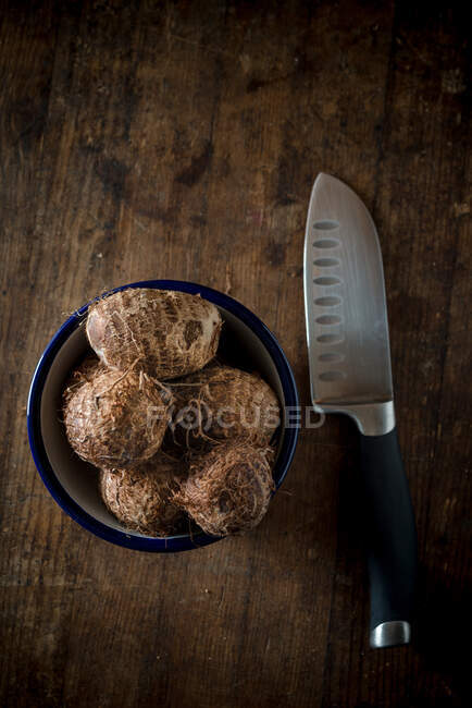 From above of ripe tropical eddo vegetables in ceramic bowl placed on wooden table near sharp knife — Stock Photo