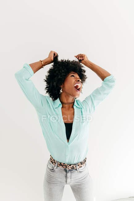 Jeune femme afro-américaine ludique en tenue tendance s'amusant à toucher les cheveux afro en regardant vers le haut sur fond blanc — Photo de stock