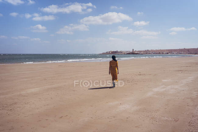 Unrecognizable distant female traveler in yellow dress standing on sandy beach with calm clear water and enjoying sunny day while spending summer holidays in Oman — Stock Photo