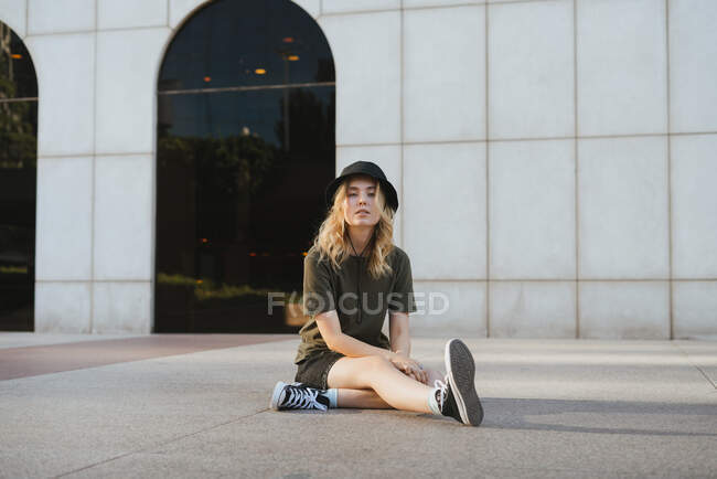 Young blond female in casual clothes and gumshoes sitting with crossed legs on walkway while looking at camera in town — Stock Photo