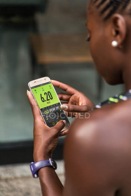 Irreconocible mujer afroamericana navegando aplicación de fitness en el teléfono inteligente mientras está de pie en el fondo borroso de la calle de la ciudad durante el entrenamiento al aire libre - foto de stock
