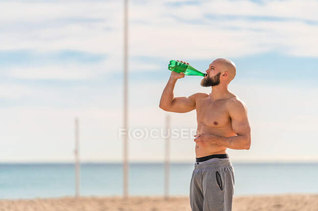 Vue latérale de l'homme barbu torse nu satisfaisant la soif après l'entraînement eau potable de la bouteille debout sur la plage de sable regardant loin — Photo de stock