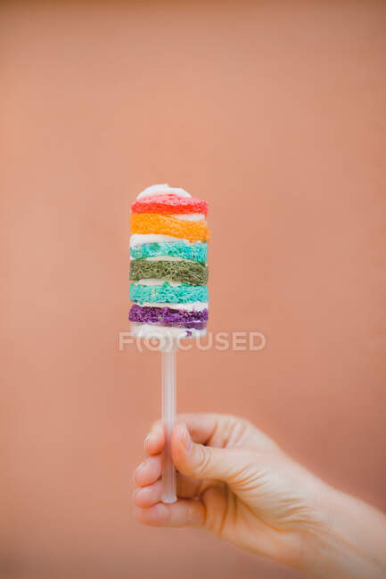Hand holding baked sweet rainbow cake with colorful cute design on brown background — Stock Photo