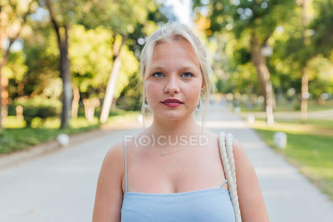 Feminino com cabelo loiro em pé no parque de verão no dia ensolarado e olhando para a câmera — Fotografia de Stock