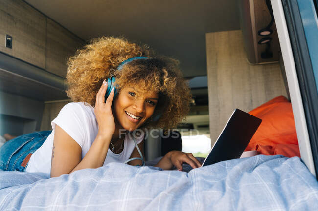 Fröhliche Afroamerikanerin lächelt und hört Musik über Kopfhörer, während sie im Wohnwagen auf dem Bett liegt und auf dem Laptop in den sozialen Medien surft — Stockfoto