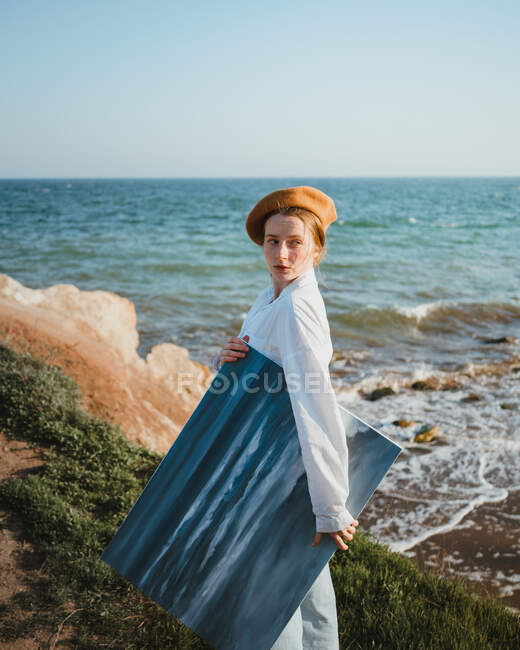 Вид сбоку молодой художницы в стильном наряде и шляпе, прогуливающейся возле песчаного пляжа волнистого моря с картиной в руке — стоковое фото