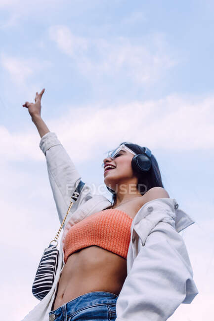 Low angle of smiling female enjoying music in wireless headphones and raising arm up with victory sign — Stock Photo