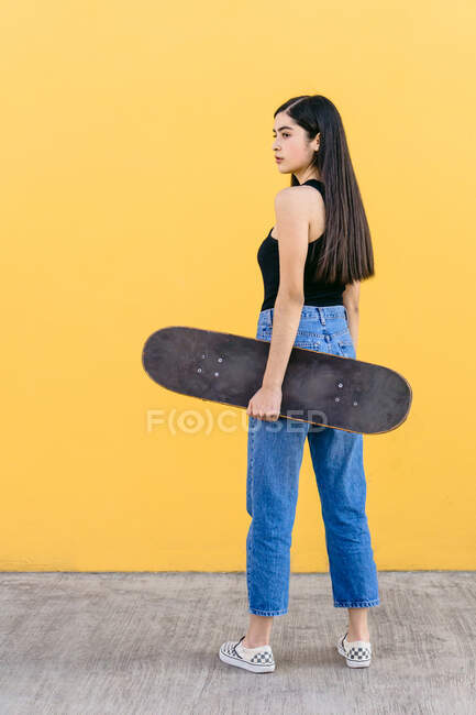 Vista lateral de la joven patinadora femenina con monopatín de pie mirando hacia otro lado en la pasarela con colorida pared amarilla en el fondo durante el día - foto de stock