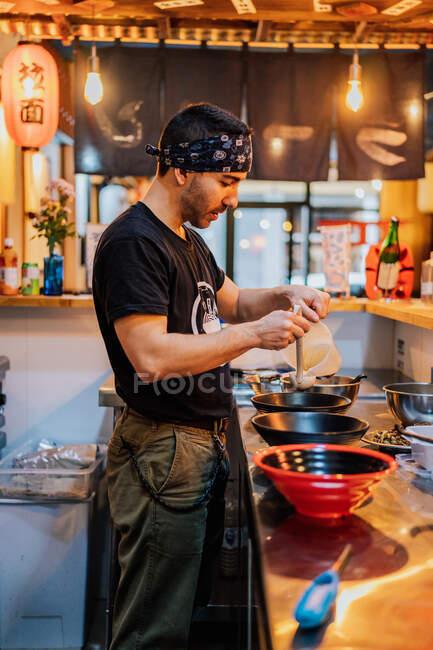 Вид сбоку на повара-мужчину в черной униформе и бандане, готовящего азиатское блюдо под названием рамен в современном кафе — стоковое фото