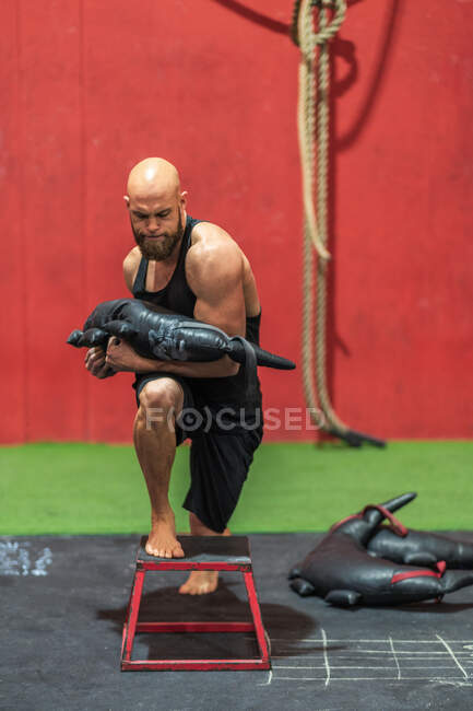 Esportista poderoso com saco pesado pisando e lunging nas fezes durante o treinamento funcional no ginásio moderno — Fotografia de Stock