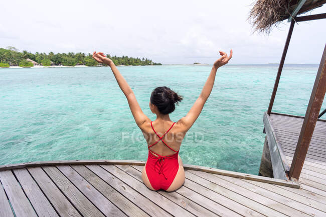 Vue arrière de la femelle méconnaissable en maillot de bain assis sur une jetée en bois relaxant aux Maldives — Photo de stock