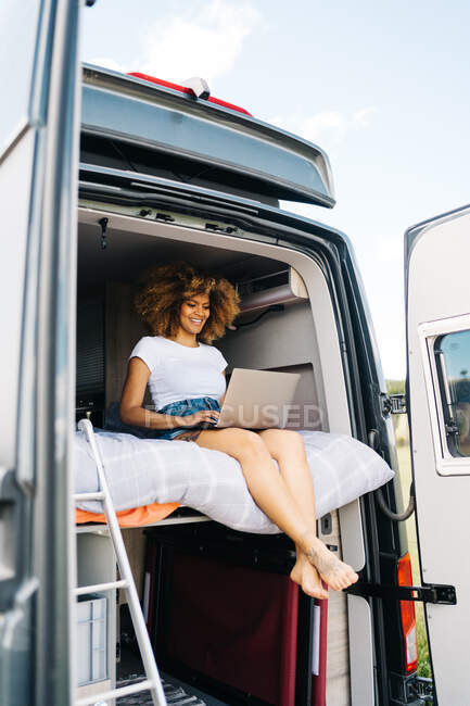 Jeune voyageuse afro-américaine avec des cheveux bouclés regarder film sur ordinateur portable tout en se reposant à l'intérieur du camping-car pendant les vacances d'été — Photo de stock