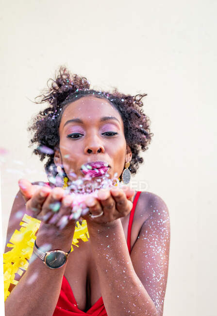 Giovane femmina etnica con acconciatura afro in accessori che soffiano su petali floreali mentre guarda la fotocamera su sfondo chiaro — Foto stock