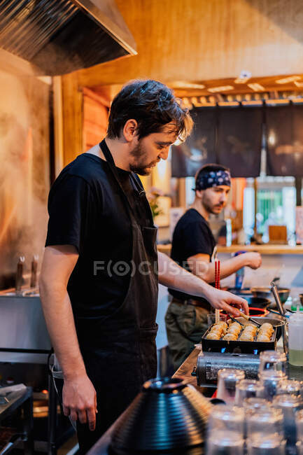 Вид сбоку на повара-мужчину в черной униформе и бандане, готовящего азиатское блюдо под названием рамен в современном кафе — стоковое фото