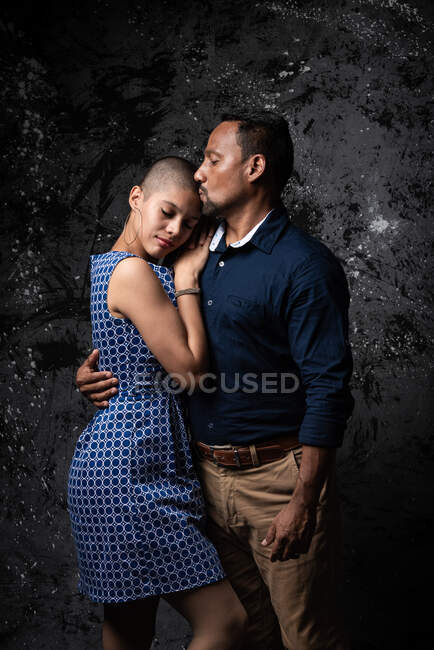 Tender homem étnico beijando mulher com os olhos fechados na cabeça no fundo escuro no estúdio — Fotografia de Stock