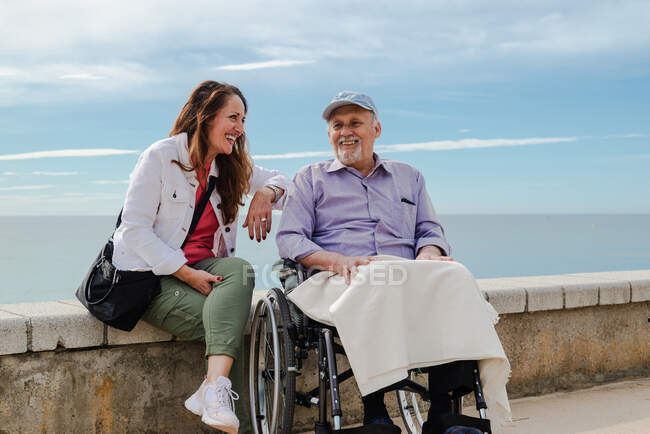 Довольна взрослая дочь и пожилой отец в инвалидной коляске охлаждения на набережной против моря вместе летом — стоковое фото
