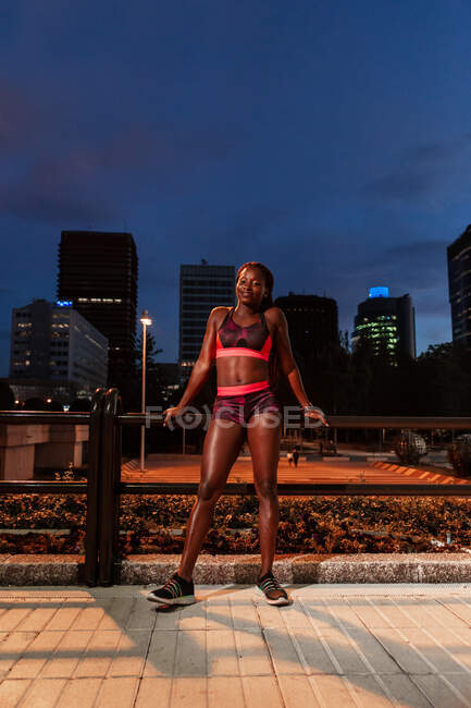 Fröhliche ethnische Sportlerin lehnt am Geländer und blickt nachts auf der Straße in die Kamera — Stockfoto