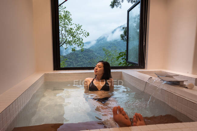 Расслабленная молодая этническая леди в купальниках, лежащая в японской ванне Онсен в спа-курорте рядом с окном с видом на горы и зеленые деревья — стоковое фото