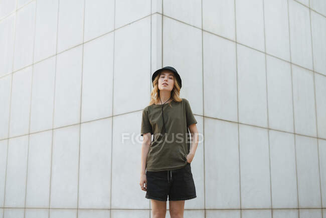 Jeune femme en vêtements décontractés regardant la caméra contre le mur de béton du bâtiment moderne sur la chaussée urbaine en journée — Photo de stock