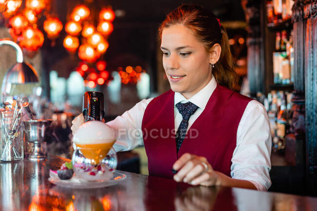 Abile giovane barista femminile utilizzando pistola fumo sapore bluster mentre guarnire cocktail al bancone del bar — Foto stock