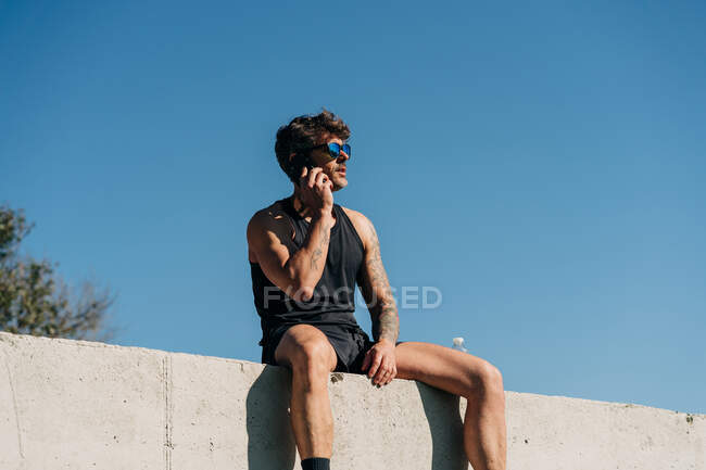 Ajuste atleta masculino en ropa deportiva hablando en el teléfono celular mientras descansa en la cerca después del entrenamiento - foto de stock