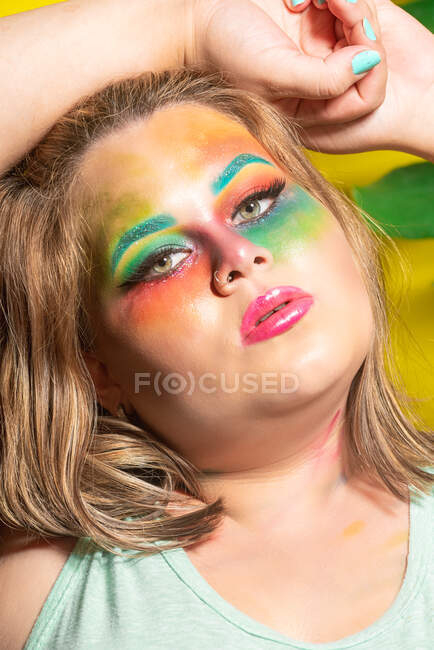 Jeune modèle féminin dodu avec maquillage créatif coloré touchant la tête et regardant la caméra sur fond jaune — Photo de stock