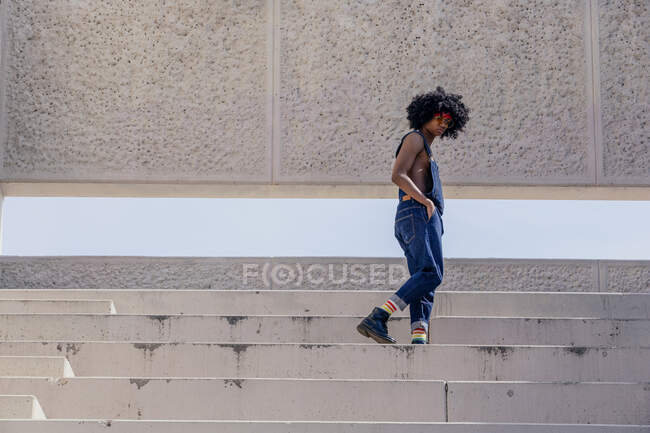 Side view of African American male in denim overalls standing with hand in pocket on stairs while looking down — Stock Photo