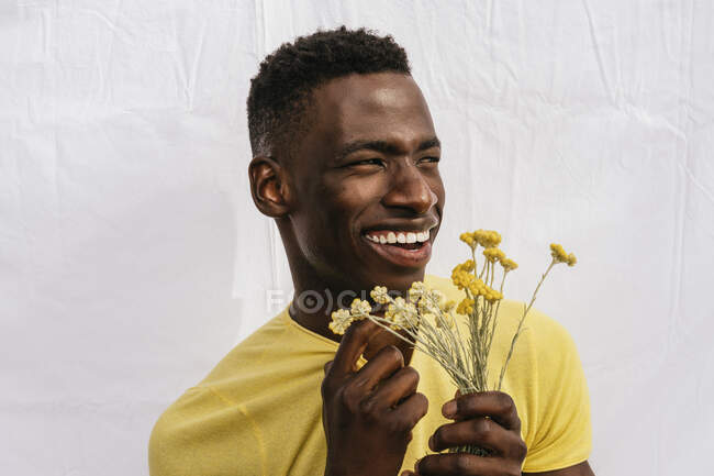 Joyeux mâle afro-américain avec bouquet de fleurs sauvages jaunes regardant loin sur fond blanc — Photo de stock
