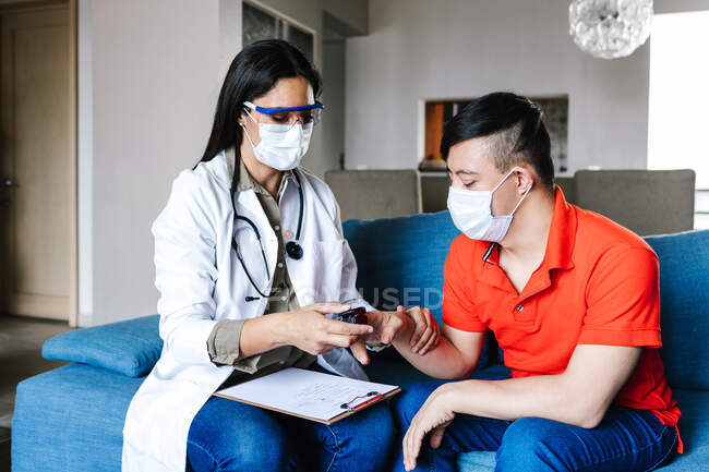 Médico femenino serio en máscara protectora usando oxímetro de pulso para adolescente latino durante cita en casa - foto de stock