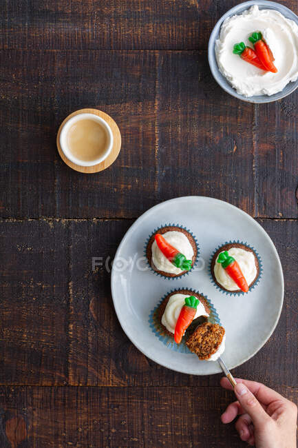 Vista dall'alto di persona ritagliata irriconoscibile mano mangiare deliziosi cupcake vegetali con piccola decorazione dolce carota sulla parte superiore posta sul piatto sul tavolo di legno — Foto stock