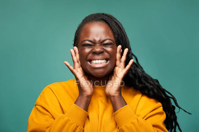 Glad African American female in yellow clothes with toothy smile and eyes closed holding face in hands against blue background — Stock Photo