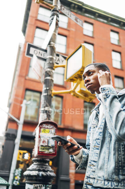 De baixo conteúdo alegre afro-americano cara em jeans jaqueta na moda surfar telefone celular moderno durante o passeio pela cidade — Fotografia de Stock