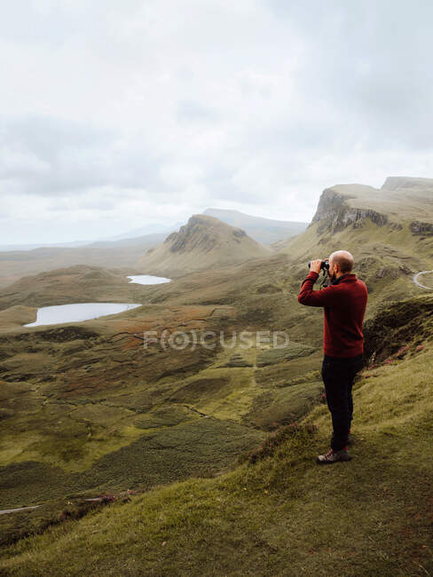Side view of bald man exploring hilly terrain through binoculars while standing on grassy slope on overcast day in UK nature — Stock Photo