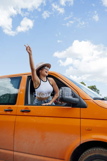 Веселая женщина-путешественница, выглядывающая из окна фургона и показывающая два пальца жестом, наслаждаясь поездкой летом — стоковое фото
