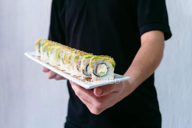 Crop anonymous male waiter standing with plate of delicious sushi rolls topped with soy sauce and sesame seeds — Stock Photo