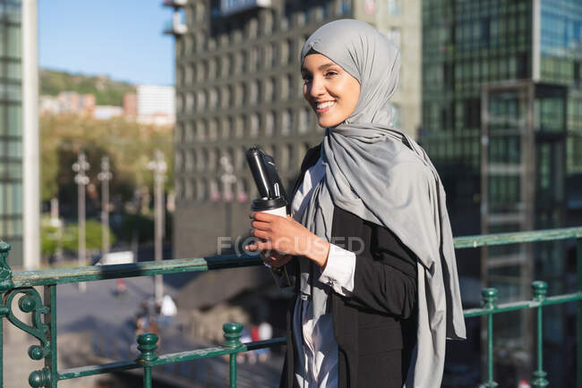 Alegre empresaria musulmana en hijab y con café para llevar de pie en la calle - foto de stock