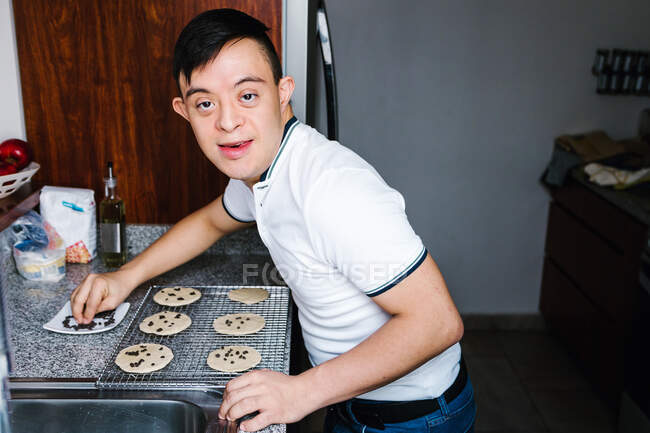 Vista laterale di allegro ragazzo latino adolescente con sindrome di Down decorare biscotti crudi con gocce di cioccolato durante la cottura in cucina a casa — Foto stock