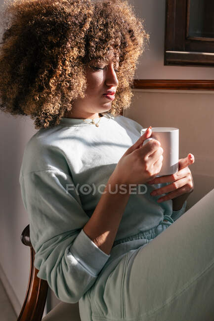 Afro-americana com cabelo encaracolado sentado no sofá e bebendo bebida quente em casa — Fotografia de Stock