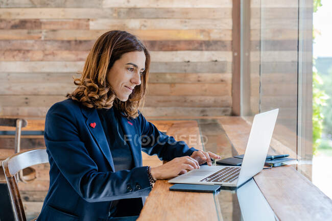 Positive erwachsene Frau in elegantem Outfit surft auf Laptop, während sie im Stuhl am Tisch neben dem Notizbuch in der hellen Cafeteria am Fenster sitzt — Stockfoto