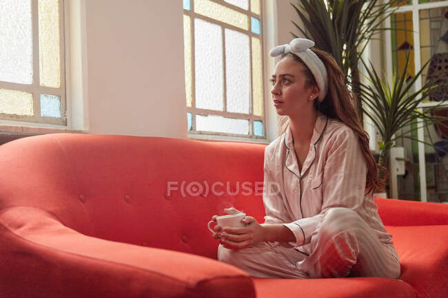 Вид збоку жінки в нічному одязі, сидячи з чашкою гарячого напою на дивані і насолоджуючись ранком вдома — стокове фото