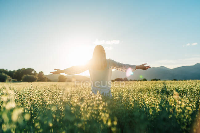 Back view of anonymous blond female in white dress raising arms showing two fingers while standing in vast summer field with blooming grass — Stock Photo